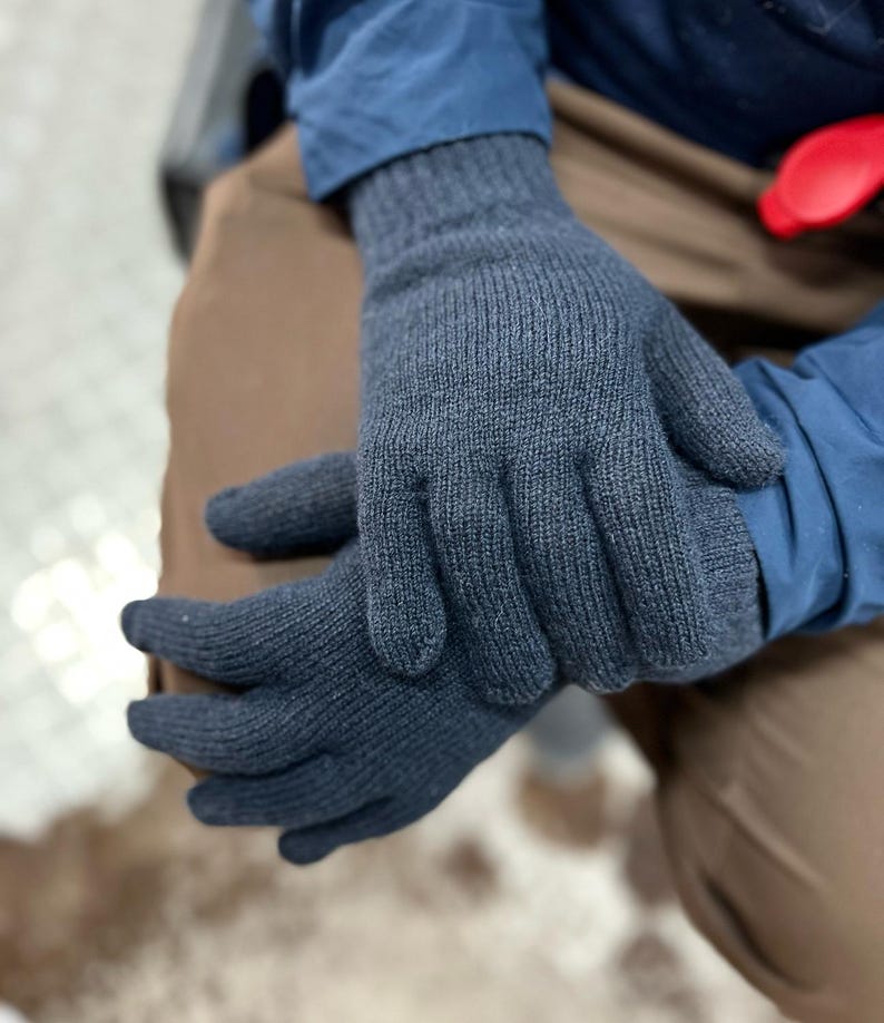 May include: A pair of dark grey knitted gloves with a ribbed cuff. The gloves are folded in half and held in the hands of a person.