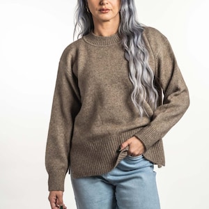 Puede incluir: Una mujer con un suéter marrón y pantalones vaqueros azul claro. Su cabello es largo y ondulado, de color azul claro.