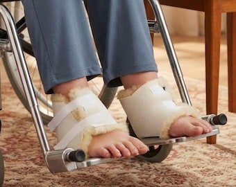 May include: A pair of off-white heel protectors with a soft, fuzzy interior lining and white hook-and-loop closure straps. These protective cushions are worn around the back of the feet to provide padding and support while resting on footrests.