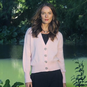 May include: A woman wearing a light pink cardigan sweater with silver buttons over a black top and black pants. The background features a green pond and trees.