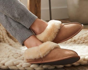 May include: Tan suede slippers featuring a plush cream-colored shearling lining and cuff. The slippers have a rounded toe design and a flat sole, providing a simple slip-on style. The exterior is a smooth, light brown material with visible stitching.