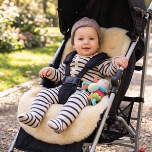 May include: A baby wearing a brown knit beanie and a long-sleeved, footed onesie with navy blue and white horizontal stripes. The baby is secured in a black stroller with a cream-colored sheepskin liner and holds a small, colorful plush toy.