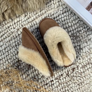 May include: A pair of brown suede slippers with a fluffy white sheepskin lining. The slippers are on a white and gray textured rug.