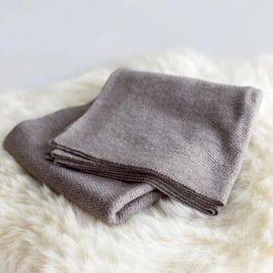 May include: Two folded grey blankets are stacked on a white, fluffy surface. The blankets have a textured appearance and are neatly folded, suggesting a cosy and comfortable theme. The neutral colour palette offers a sense of calm.