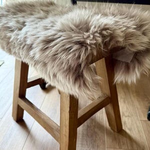 May include: A wooden stool with a light brown, shaggy faux fur seat cover. The stool has a simple, rustic design with four legs and a square seat. The fur is a mix of light brown and gray tones, creating a soft, textured appearance.
