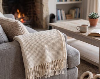 May include: A cream-colored waffle-knit throw blanket with long fringe trim draped over the arm of a gray upholstered sofa. The blanket features a textured grid pattern and is positioned in a living room setting with a fireplace and coffee table.