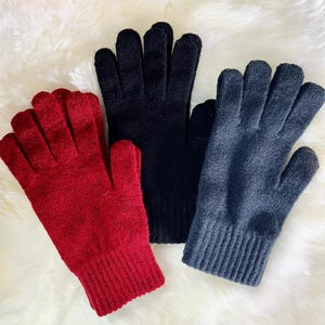 May include: Three pairs of knitted gloves in red, black and grey. The gloves are folded and laid out on a white fluffy surface.