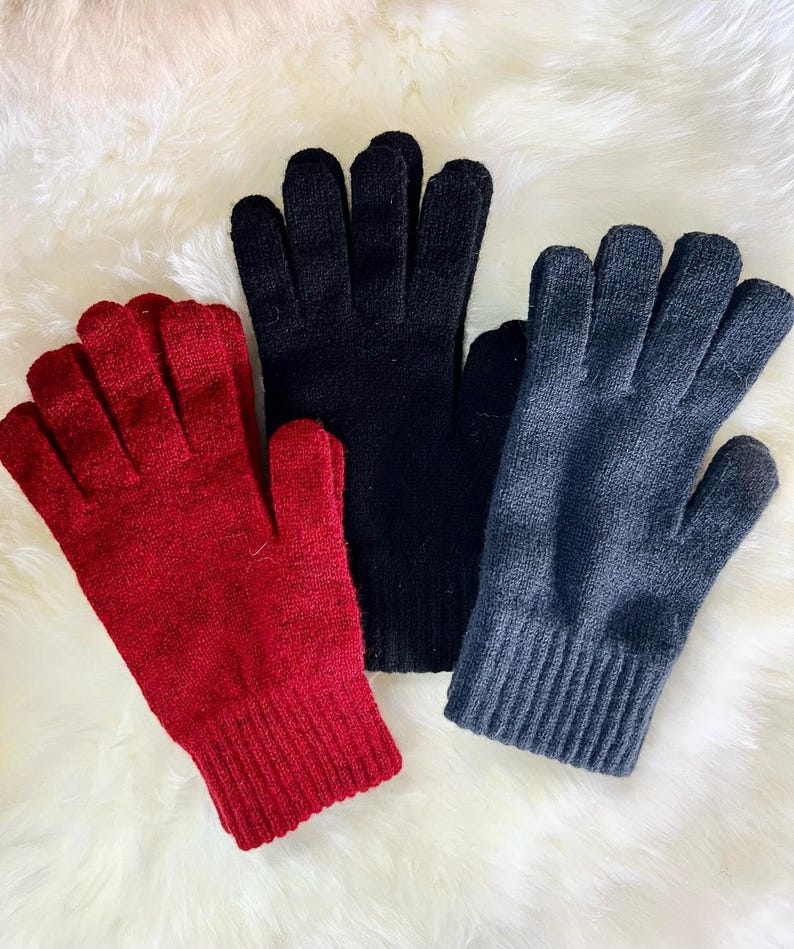 May include: Three pairs of knitted gloves in red, black and grey. The gloves are folded and laid out on a white fluffy surface.