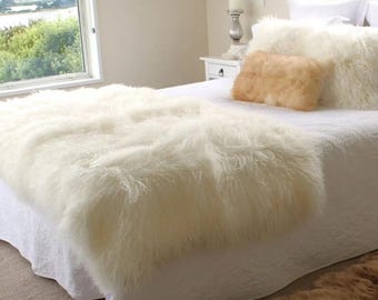 May include: A white long-haired sheepskin throw blanket draped over a bed with a white quilted coverlet. A rectangular tan sheepskin pillow sits against two white pillows. A separate tan sheepskin rug is positioned on the floor near the bed.