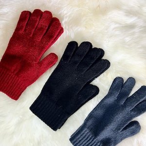 May include: Three pairs of knitted gloves in red, black and grey. The gloves are laid out on a white fluffy surface.