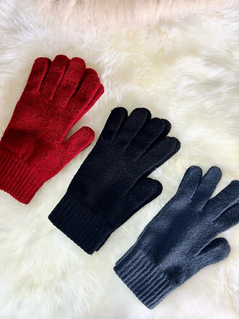 May include: Three pairs of knitted gloves in red, black and grey. The gloves are laid out on a white fluffy surface.