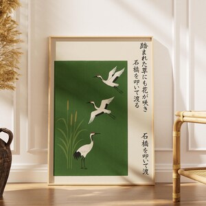 May include: A framed art print featuring three white cranes in flight against a green background. The artwork includes Japanese text and stylized reeds. The frame is a light wood color, and the overall aesthetic is minimalist and nature-inspired.