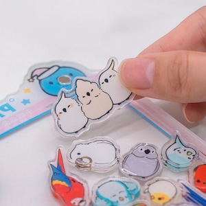 May include: A selection of clear acrylic stickers with cartoon bird designs. One sticker shows three birds, two white and one beige, held by a hand. Other stickers include a blue bomb with a Santa hat, a parrot, and other bird designs.
