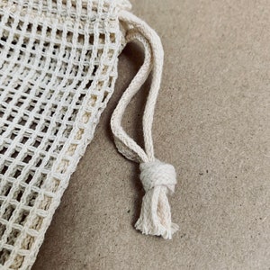 May include: A close-up of a white mesh produce bag with a drawstring closure. The bag is made of a fine, woven material and has a natural, unbleached color.