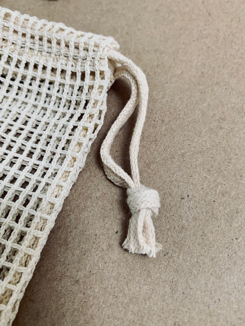 May include: A close-up of a white mesh produce bag with a drawstring closure. The bag is made of a fine, woven material and has a natural, unbleached color.