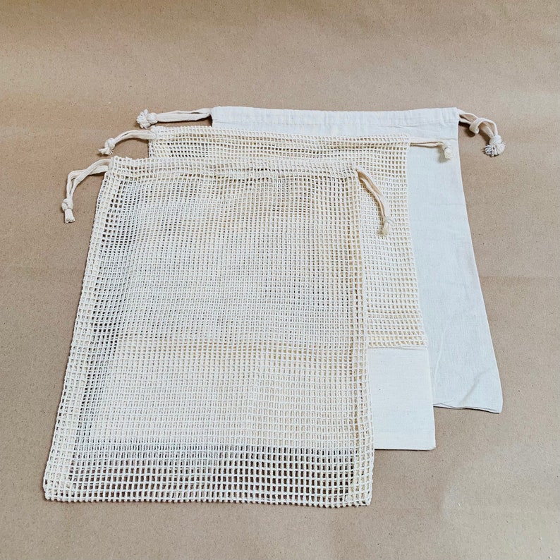May include: Three reusable produce bags made of white mesh fabric with drawstring closures. The bags are stacked on top of each other on a brown background.