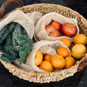 May include: A wicker basket filled with produce, including apples, oranges, and leafy greens. The produce is stored in reusable mesh bags.