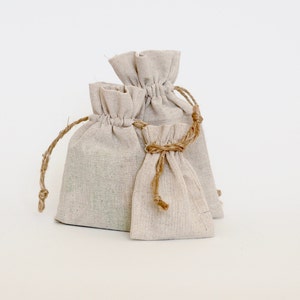 May include: Three beige linen drawstring bags of varying sizes. The bags have a natural, textured appearance and are tied with twine. The bags are arranged against a white background, suggesting a focus on the product itself.