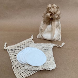 May include: Reusable white cotton rounds and mesh bags. One bag is filled with rounds and tied with a burlap bow. Two additional bags with drawstring closures are filled with rounds. The bags are beige and the background is a light brown.
