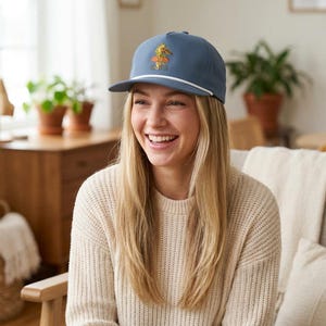 May include: A slate blue baseball cap featuring a white cord trim along the brim and an embroidered graphic of a green frog sitting on a mushroom with the text EMPS printed in gold lettering centered on the front panel of the hat.