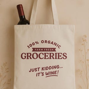 May include: Cream-colored tote bag with burgundy text reading "100% Organic Groceries, Just Kidding... It's Wine!" A bottle of red wine is partially visible inside the bag. The bag has long handles.