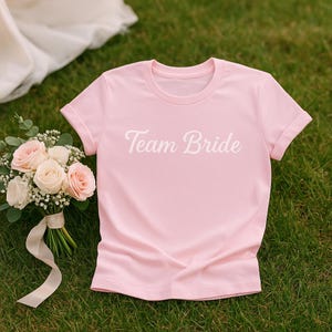 May include: A light pink t-shirt with the text "Team Bride" in white script. The shirt is laying on green grass next to a bouquet of roses and greenery. The bouquet is tied with a white ribbon.