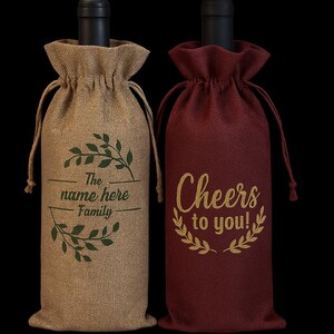 May include: Two wine bottle bags with drawstring closures. The left bag is tan burlap with a green leaf wreath design and the text "The name here Family". The right bag is burgundy burlap with a gold leaf wreath design and the text "Cheers to you!".