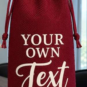 May include: A deep red burlap drawstring wine bottle bag featuring the white text YOUR OWN Text printed on the front. The bag has a textured woven fabric finish and matching red cord drawstrings that cinch at the top to secure the contents.