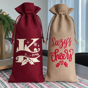 May include: Two tall drawstring wine bags made of textured fabric. The left bag is deep red with a cream-colored graphic featuring a large letter K, the word family, pine trees, a snowflake, and holly. The right bag is tan with red text reading Suzy's cheers above a holly icon.
