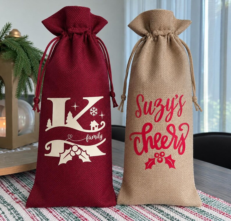 May include: Two tall drawstring wine bags made of textured fabric. The left bag is deep red with a cream-colored graphic featuring a large letter K, the word family, pine trees, a snowflake, and holly. The right bag is tan with red text reading Suzy's cheers above a holly icon.