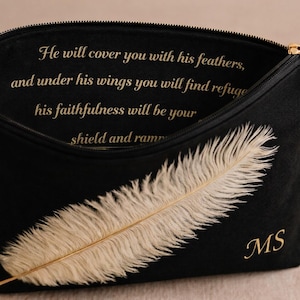 May include: Black zippered pouch with a gold-colored zipper and tassel. The pouch features a large, cream-colored feather design and the initials "MS" in gold. Text in gold lettering reads, "He will cover you with his feathers..."