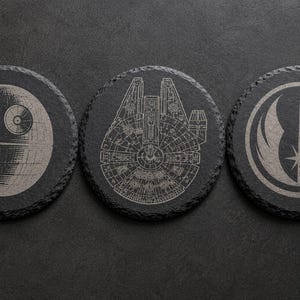 May include: Three circular black slate coasters with rough, textured edges. Each coaster features a different laser-engraved design: a Death Star, a Millennium Falcon, and a Jedi Order symbol. The engravings are light gray against the dark stone.