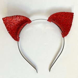 May include: A red glitter cat ear headband. The ears are a vibrant red and covered in sparkling glitter. The headband is a thin, silver-coloured metal. The ears are attached to the headband with a small amount of glue.