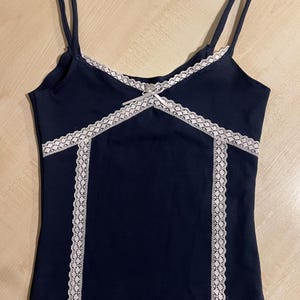 Può includere: Canotta blu navy con sottili spalline. La parte superiore presenta finiture in pizzo bianco lungo lo scollo e i lati, con un piccolo dettaglio a fiocco al centro. Un capo femminile ed elegante.
