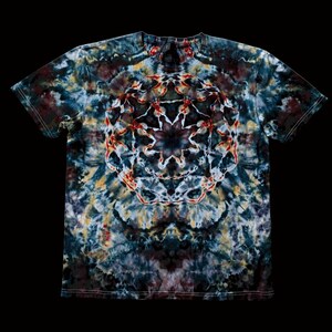 May include: A short-sleeve t-shirt featuring a tie-dye pattern in shades of dark blue, black, grey, and yellow. The center of the shirt displays a prominent, symmetrical geometric mandala design in white and red, creating a stark contrast.