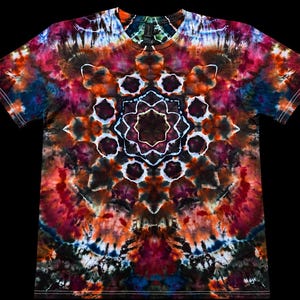 May include: A short-sleeve crew neck t-shirt featuring a complex tie-dye pattern with a central star-shaped mandala design. The garment displays a vibrant color palette of deep red, orange, dark blue, black, and white hues throughout the fabric.