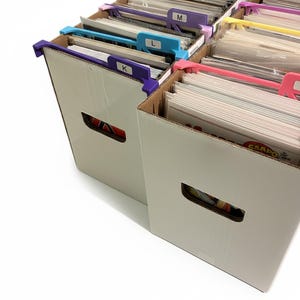 Puede incluir: Dos cajas de almacenamiento de cartón blanco llenas de cómics. Las cajas tienen asas recortadas y están etiquetadas con pestañas de colores marcadas K, L y M. Las pestañas son moradas, azules y rosas. Las cajas están sobre un fondo blanco.