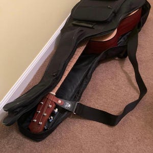 May include: Acoustic guitar in a black case. The guitar has a dark red body and a wooden neck. The case has a shoulder strap and a small pocket. The guitar is partially visible, suggesting it's being stored or transported.