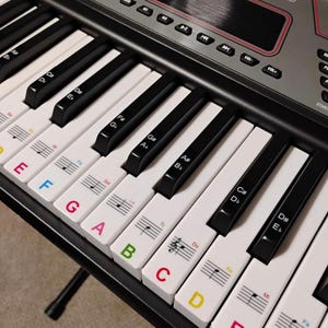 May include: A black and silver electronic keyboard with white and black keys. The keys have colorful letter and musical note stickers. The keyboard has various function buttons and a display screen.