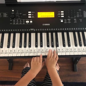 May include: Black Yamaha keyboard with white and black keys, labeled with musical notes. The display reads "Grand Piano." Hands are positioned on the keys, with feet and striped pants visible below. The keyboard is on a black stand.