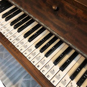 Könnte beinhalten: Nahaufnahme einer Vintage-Klaviertastatur mit schwarzen und weißen Tasten. Die Tasten sind mit Noten und Symbolen beschriftet. Der Holzrahmen hat eine dunkelbraune Oberfläche. Die Klaviertasten sind mit den Noten A bis G beschriftet.