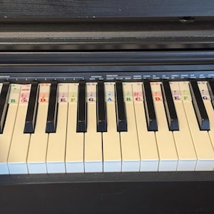 May include: Close-up of a black digital piano keyboard with white and black keys. Colorful stickers label the notes with letters and symbols. The piano is set against a dark wooden background.