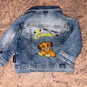 May include: A light blue denim jacket featuring a Simba design on the back. The design includes yellow text, a cartoon lion, and black birds. The jacket has a button closure and two front pockets.