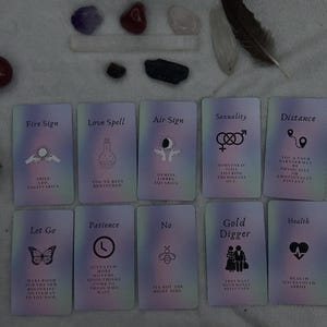 Pode incluir: Uma variedade de cartas de tarô com vários símbolos e texto, incluindo "Fire Sign", "Love Spell" e "Air Sign". As cartas são dispostas com cristais, um bastão de fumo e penas. As cartas têm um fundo macio e iridescente.