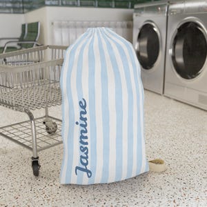 May include: A large laundry bag with vertical light blue and white stripes. The name "Jasmine" is printed vertically in a cursive font on the side of the bag. The bag is sitting on a speckled floor near a laundry cart.