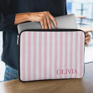May include: A laptop sleeve with a pink and white striped pattern. The sleeve has a black border and a zipper closure. The name "OLIVIA" is printed in a matching pink font on the front of the sleeve.