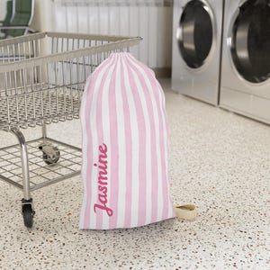Pode incluir: Uma bolsa de roupa suja listrada em rosa e branco com o nome "Jasmine" em fonte cursiva. A bolsa está em pé ao lado de um carrinho de compras de metal em uma lavanderia.