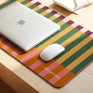 May include: A rectangular desk mat featuring a repeating vertical striped pattern in shades of mustard yellow, forest green, pink, and maroon. The text "you got this." is printed in white in the bottom right corner of the mat.
