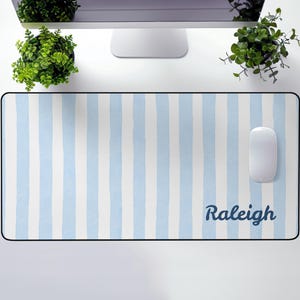May include: A rectangular desk mat featuring a light blue and white striped design. The name "Raleigh" is written in dark blue script. A white computer mouse is positioned on the right side of the mat.