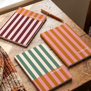 Puede incluir: Tres cuadernos con cubiertas a rayas en tonos rosa, marrón y verde, con los textos "Half-Baked Thoughts", "Ideas I'll Never Use" y "Brilliant Eventually". Un bolígrafo de madera descansa sobre la mesa.